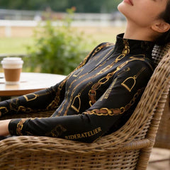 Model wearing the RIDER ATELIER Black Gold Chain & Stirrup Print Riding Top, relaxing at an outdoor café table with a coffee cup
