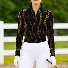 Model wearing the RIDER ATELIER Black Gold Chain & Stirrup Print Riding Top at a sunlit pasture, holding a riding crop

