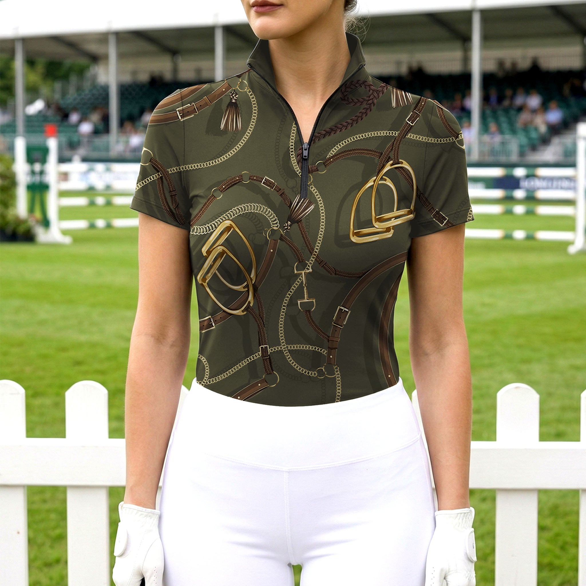 Model wearing the RIDER ATELIER Olive Equestrian Tack Print Short-Sleeve Riding Top at an outdoor equestrian arena, with a white fence in the background
