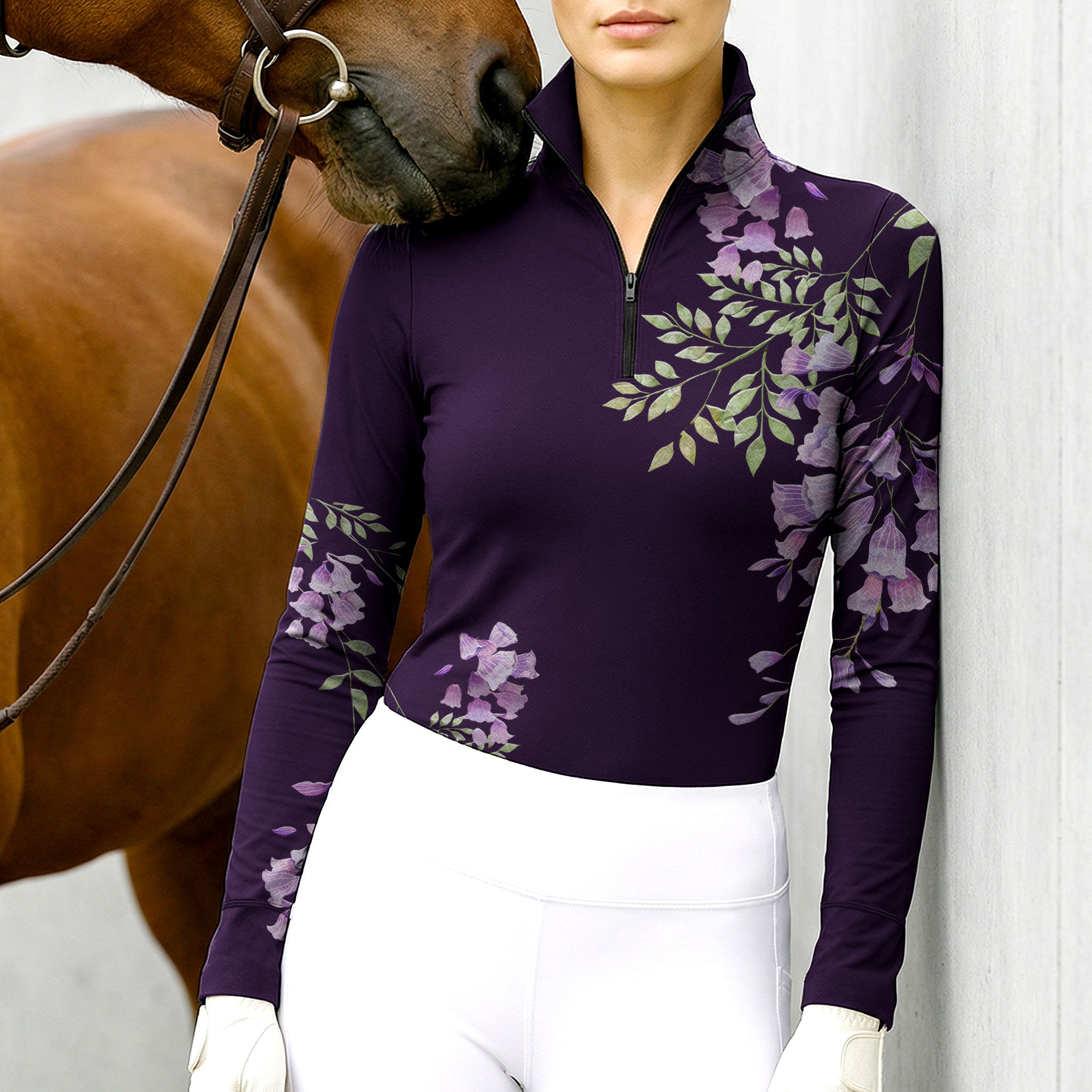 Woman wearing the RIDER ATELIER Purple Wisteria Floral Print Long-Sleeve Quarter-Zip Riding Top leaning against a stable wall, with a chestnut horse nuzzling her shoulder, highlighting the print’s soft green foliage and purple blooms up close

