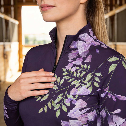 Closeup shot of the RIDER ATELIER Purple Wisteria Floral Print Long-Sleeve Quarter-Zip Riding Top in a sunlit barn, showcasing the delicate wisteria print cascading from the shoulder, with the quarter-zip collar in view
