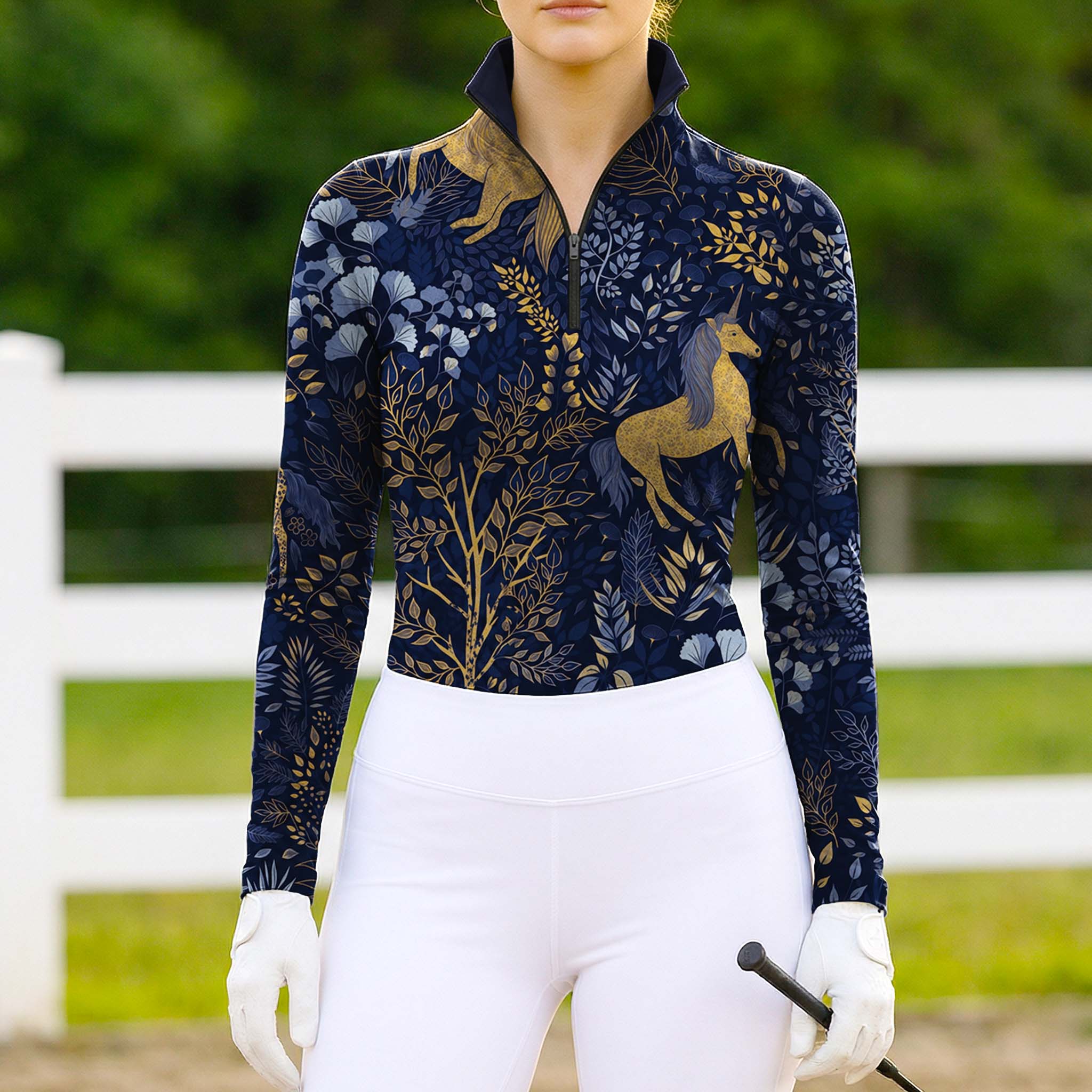 Model wearing the RIDER ATELIER Midnight Unicorn & Forest Print Riding Top at a sunlit pasture, holding a riding crop
