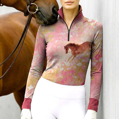 Woman wearing the RIDER ATELIER Watercolor Floral Horse Print Long-Sleeve Quarter-Zip Riding Top leaning against a stable wall, with a chestnut horse nuzzling her shoulder, highlighting the print’s delicate floral details and soft gradient tones
