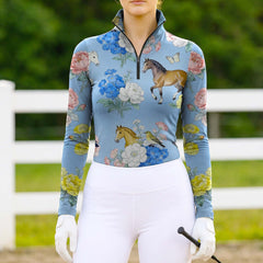 Model wearing the RIDER ATELIER Watercolor Floral Horse Print Riding Top at a sunlit pasture, holding a riding crop

