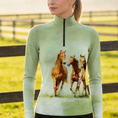 Woman wearing the RIDER ATELIER Sage Green Watercolor Galloping Horses Print Long-Sleeve Quarter-Zip Riding Top at sunset in a sunlit pasture, showcasing the print’s soft watercolor details in warm golden light
