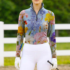 Woman wearing the RIDER ATELIER Watercolor Iris & Peony Print Light Blue Long-Sleeve Quarter-Zip Riding Top at a sunlit pasture with a white fence, holding a riding crop, showing the print’s vibrant colors in natural daylight
