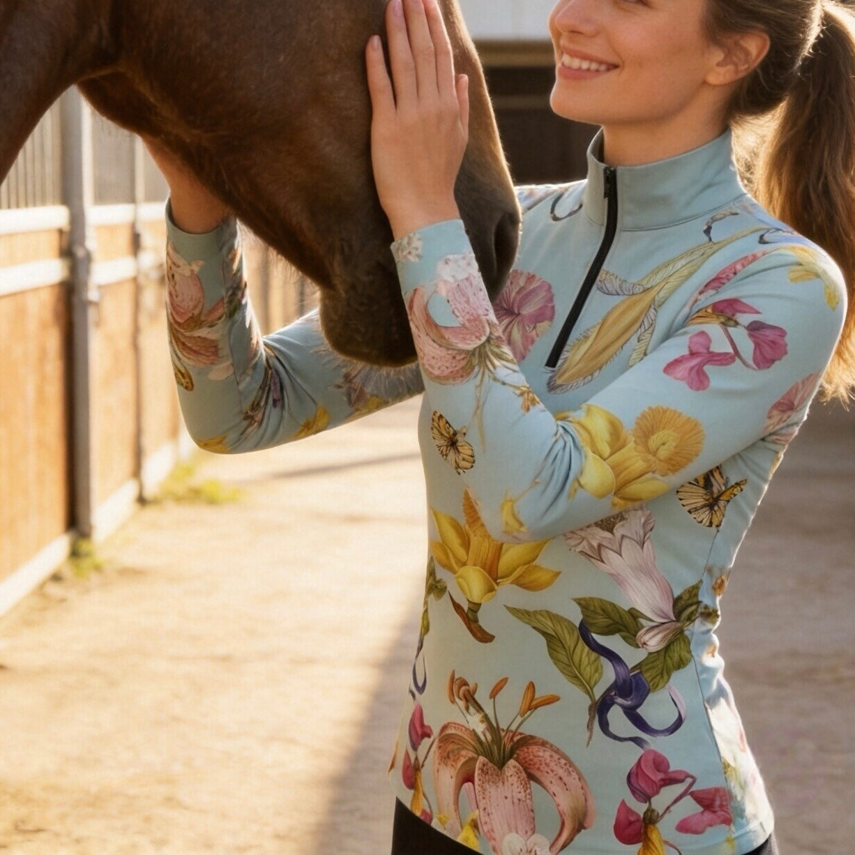 Close-up model shot petting brown horse wearing sage mint lily butterfly floral long sleeve riding top in sunlit dirt stable arena
