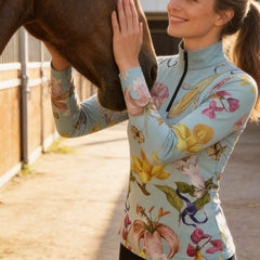 Close-up model shot petting brown horse wearing sage mint lily butterfly floral long sleeve riding top in sunlit dirt stable arena
