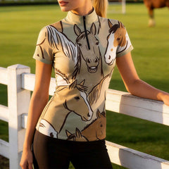 Model leaning on white wooden pasture fence at golden hour wearing sage green hand drawn multi horse portrait short sleeve riding top, brown horse blurred in background