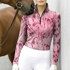 Barn stable lifestyle shot of female equestrian beside brown horse wearing blush‑pink sketch‑galloping‑horse print long‑sleeve quarter‑zip riding top