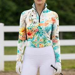 Full-body model shot wearing lush hibiscus palm hope cross horse long sleeve top at sunlit green pasture white wooden fence, holding riding crop
