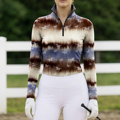 Model leaning on white wooden pasture fence wearing rustic ranch tie-dye horse silhouette long sleeve riding top in sunlit green grass field
