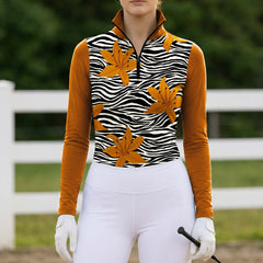 Full-body model shot wearing contrast burnt orange sleeve zebra floral riding top at an outdoor equestrian jumping arena holding a riding crop

