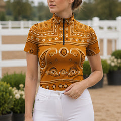 Garden paddock lifestyle shot of female rider wearing burnt‑orange retro bandana print short‑sleeve riding top with white breeches near white fencing and green shrubbery