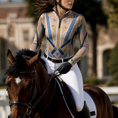 Outdoor equestrian scene shot of woman mounted on horse, riding while wearing blush‑blue art‑deco geometric‑star print long‑sleeve quarter‑zip riding top with full riding gear
