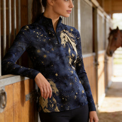 Indoor barn stall lifestyle shot of female rider leaning on stall partition wearing navy galaxy‑print long‑sleeve riding top with golden horse graphicsIndoor barn stall lifestyle shot of female rider leaning on stall partition wearing navy galaxy‑print long‑sleeve riding top with golden horse graphics