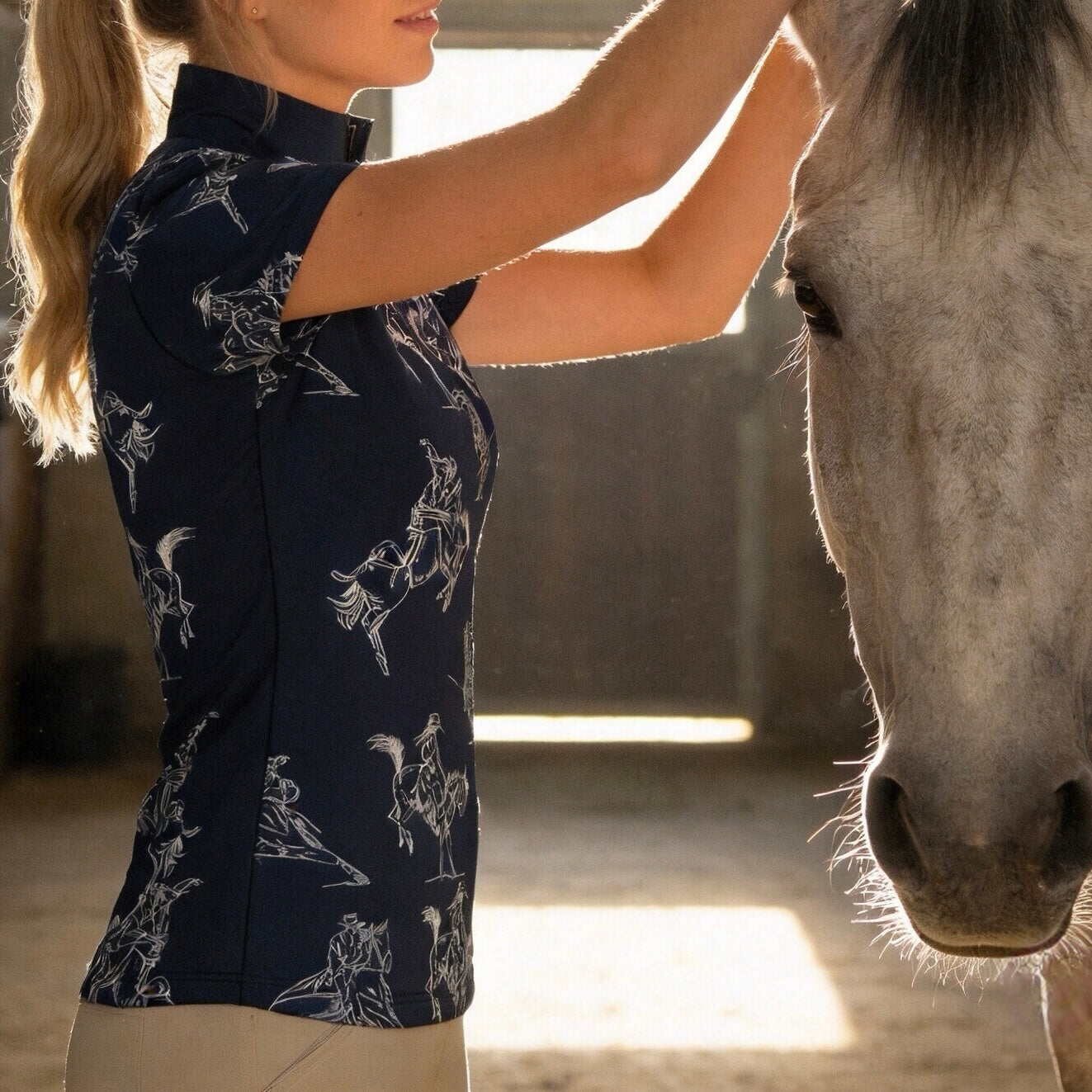 Stable indoor close‑up lifestyle shot of female equestrian wearing navy western bronco sketch‑print short‑sleeve riding top beside grey horse