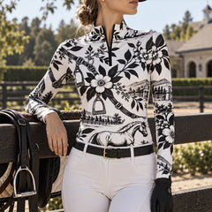 Rider in the printed top leaning on a wooden barn fence with stables visible in the background
