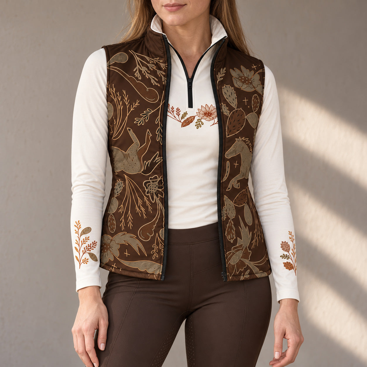 Woman wearing an open brown western horse and floral print quilted vest over a white quarter zip long sleeve top with brown riding breeches