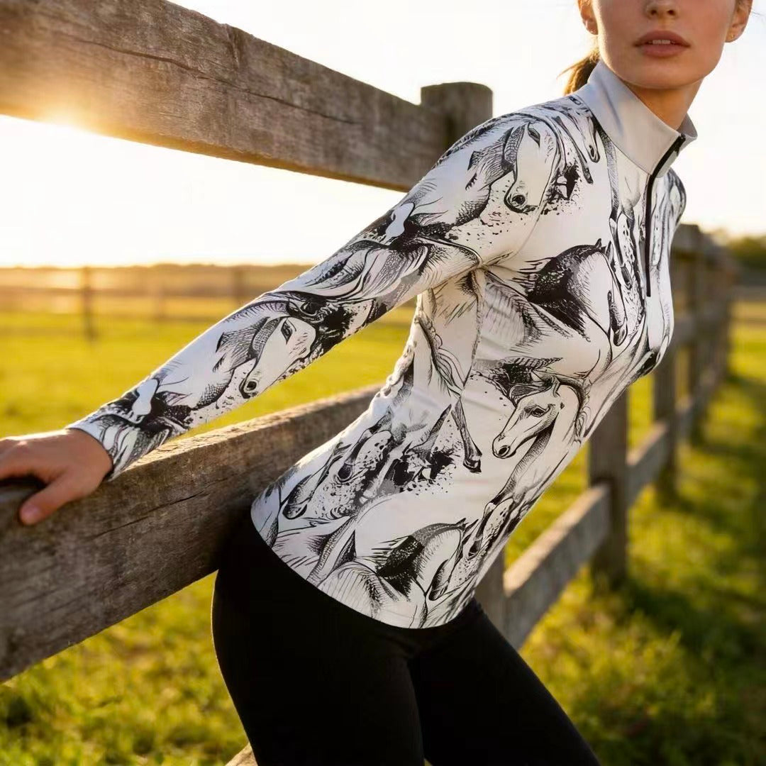 Golden‑hour pasture wooden‑fence lifestyle shot of female equestrian wearing white sketch‑galloping‑horse ink‑splash print long‑sleeve quarter‑zip riding top