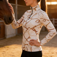 Sunlit stable arena lifestyle shot of female equestrian wearing cream bridle‑pattern long‑sleeve riding top beside brown horse
