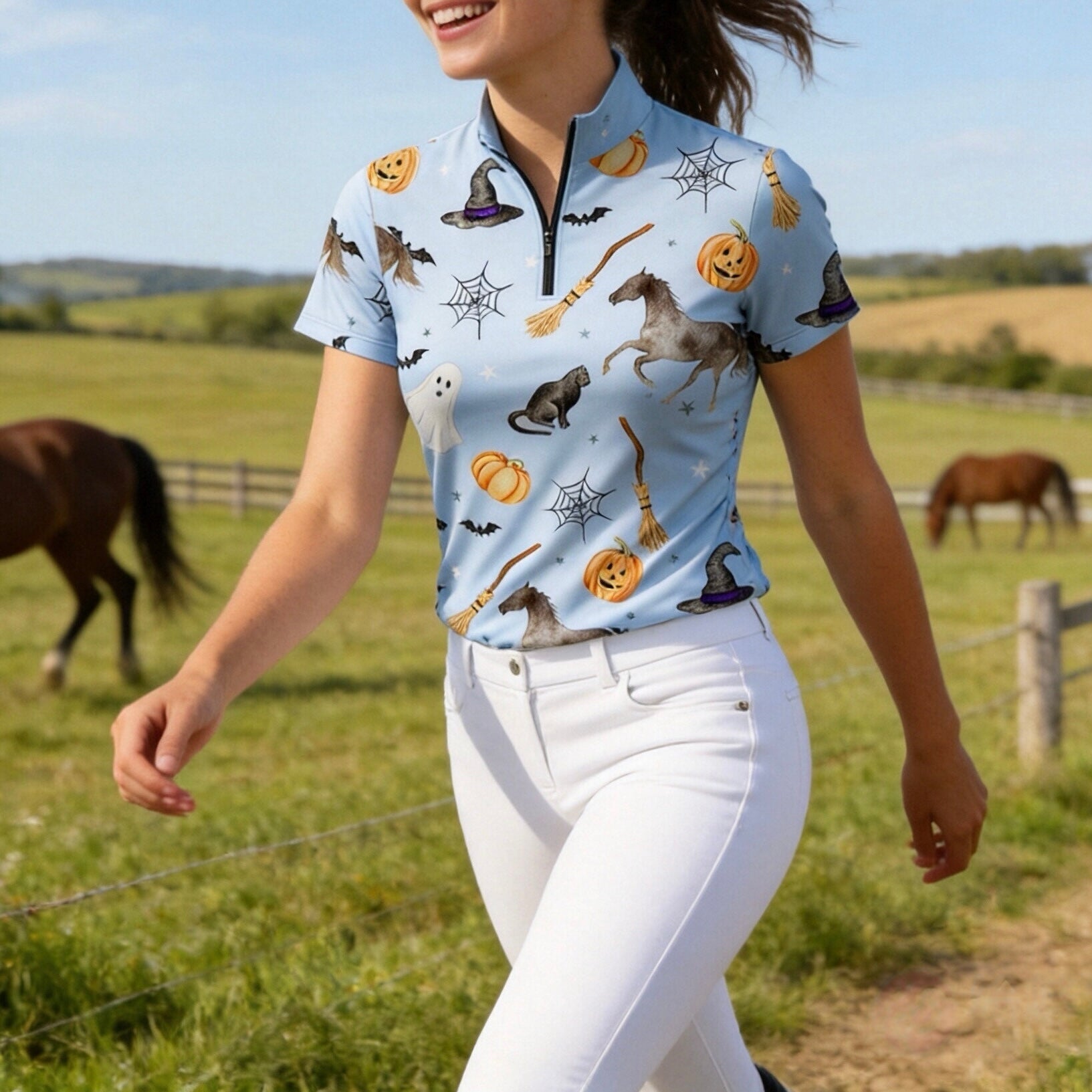 Bright daytime pasture lifestyle shot of female equestrian walking in pale‑blue short‑sleeve riding top with Halloween all‑over print