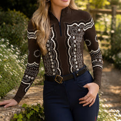 Sun‑dappled garden path lifestyle shot of female rider wearing chocolate‑brown bandana‑paisley print long‑sleeve riding top, paired with navy riding breeches and leather belt