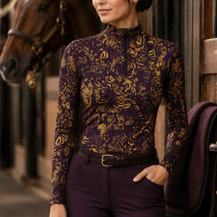 Stable yard lifestyle shot of female equestrian standing near brown horse wearing deep‑purple golden art‑print long‑sleeve riding top and purple riding breeches