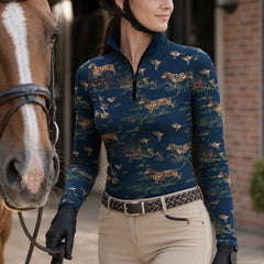 Brick stable outdoor lifestyle shot of female equestrian holding horse lead beside chestnut horse wearing navy hunting‑scene hound dog pheasant print long‑sleeve riding top with black gloves and khaki breeches