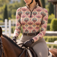 Woman wearing a light beige floral print long sleeve quarter zip riding top while seated on a brown horse and holding black reins, with a blurred green outdoor equestrian background
