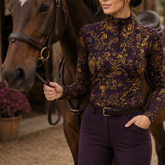 Sunny stable‑yard lifestyle shot of female equestrian holding horse reins while wearing deep‑purple golden abstract floral‑geometric doodle print long‑sleeve riding top paired with purple breeches