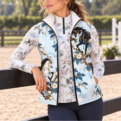 Woman wearing pale blue horse and floral print quilted vest over a printed long sleeve quarter zip top in a sandy riding arena