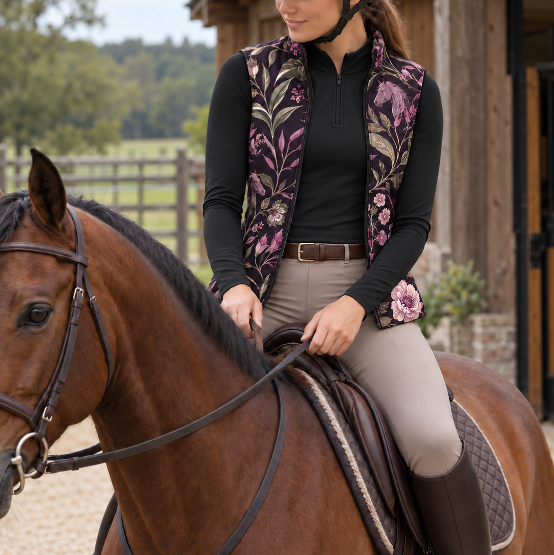 Woman wearing a dark horse and floral print reversible vest over a black long sleeve top with tan breeches and tall brown boots near a stable