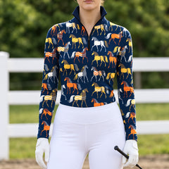 Woman wearing a navy blue horse print long sleeve quarter zip riding top with white breeches, white gloves, and a black crop in an outdoor arena with a white fence