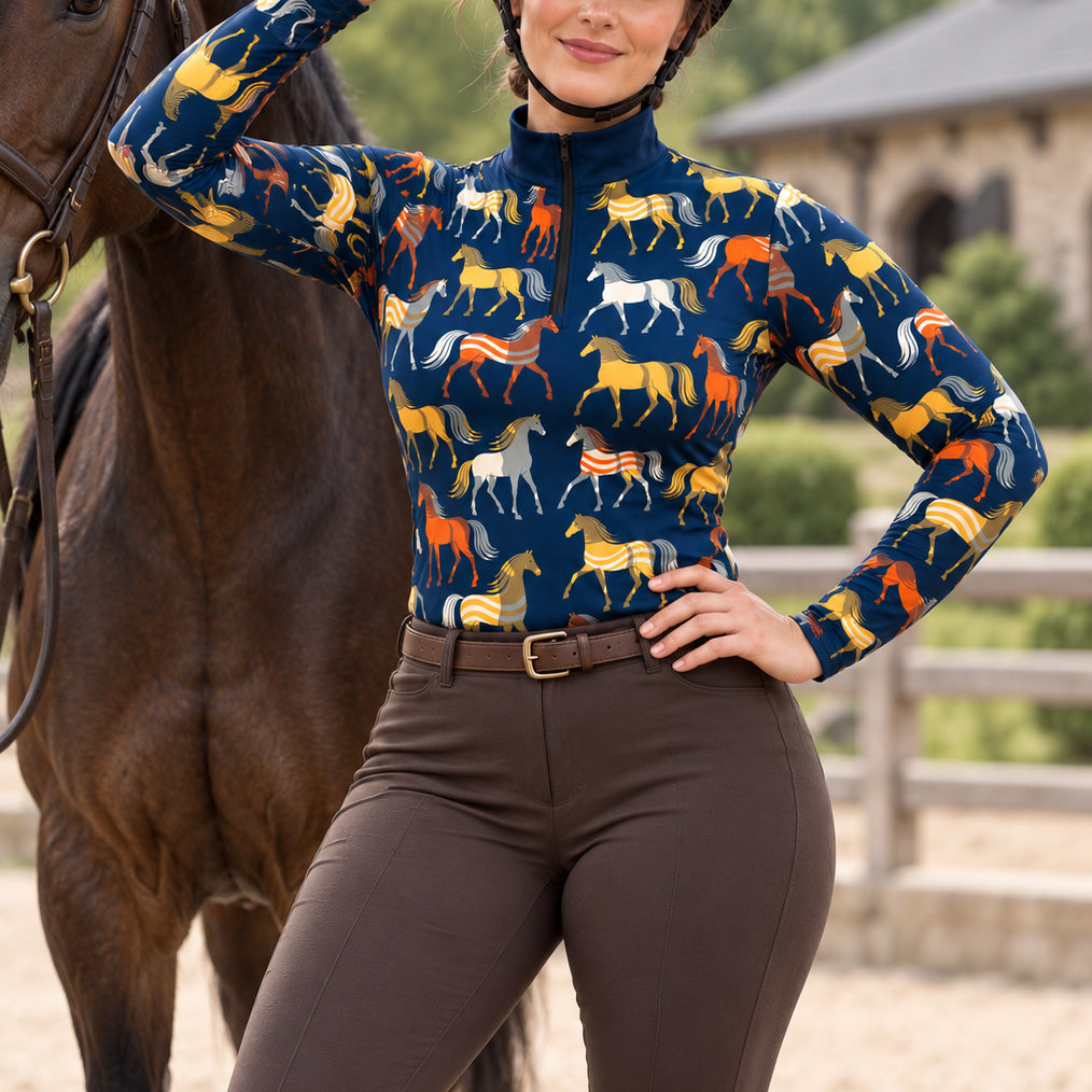 Woman wearing a navy blue horse print long sleeve quarter zip riding top with brown breeches, a brown belt, and a black helmet, next to a dark brown horse near a stone stable
