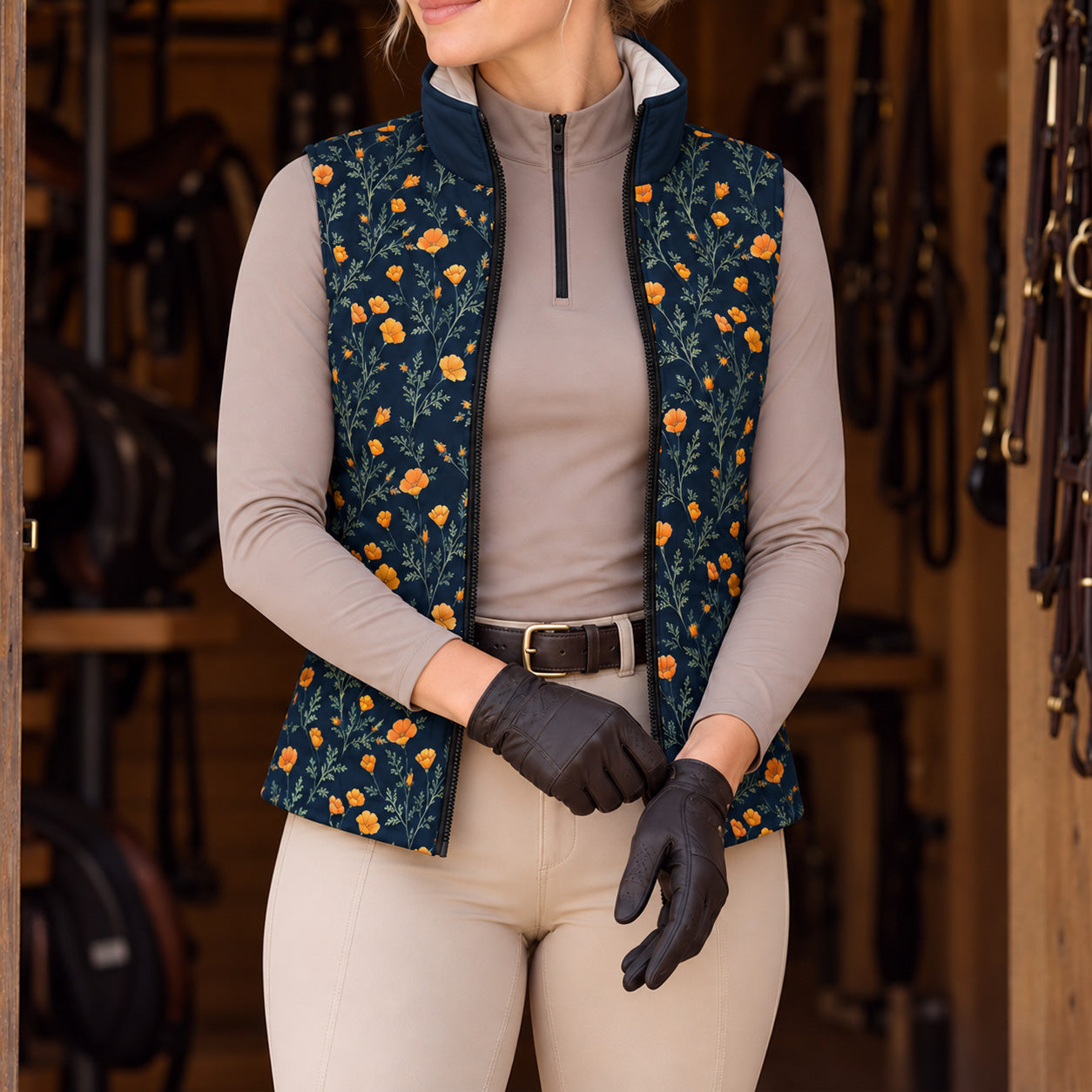 Model in a tack room wearing the navy floral print vest open over a taupe quarter zip top, beige breeches, brown leather belt, and dark brown leather riding gloves