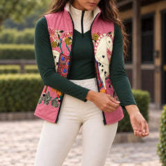 Pink printed side of reversible quilted equestrian vest worn open over a dark green long sleeve quarter zip top and white riding breeches in an outdoor barn setting