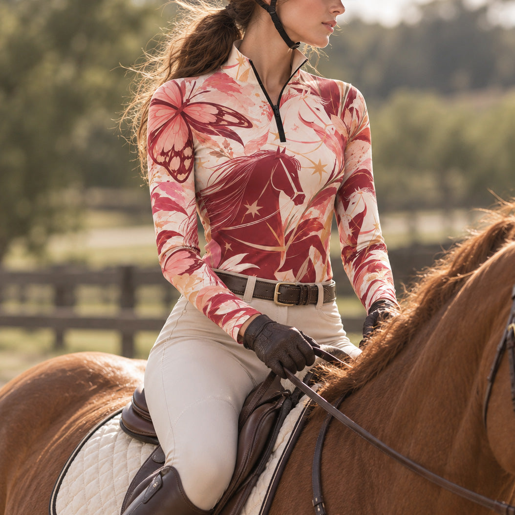 Woman wearing the cream horse and butterfly print quarter zip riding top with riding gloves and breeches in an outdoor equestrian setting