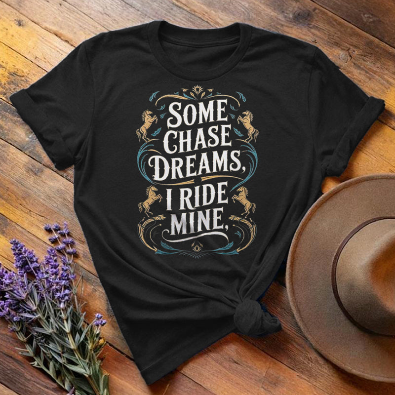 Black "Some Chase Dreams, I Ride Mine" equestrian t-shirt, flat lay on wooden background