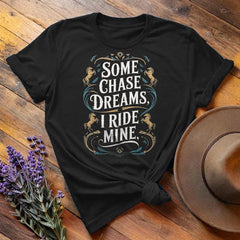 Black "Some Chase Dreams, I Ride Mine" equestrian t-shirt, flat lay on wooden background