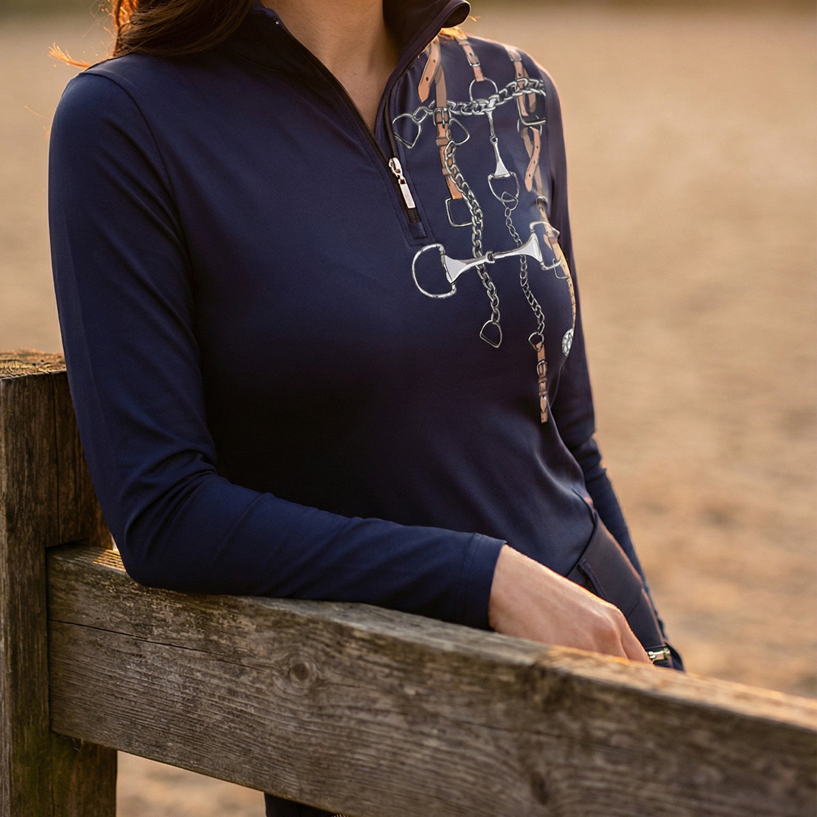 Side view of woman wearing navy equestrian tack bouquet print quarter-zip top leaning on wooden stable fence