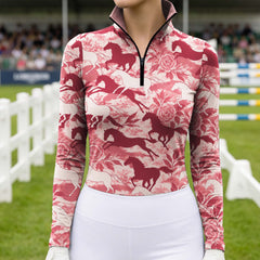 Woman wearing the light pink floral horse print quarter zip riding top at a show jumping arena with white jump obstacles and spectator stands