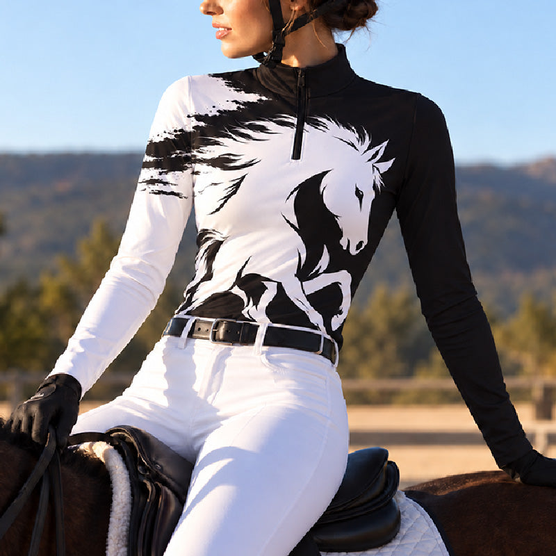 Woman wearing a black and white long sleeve quarter zip riding top with white riding breeches and a black belt in a stable setting