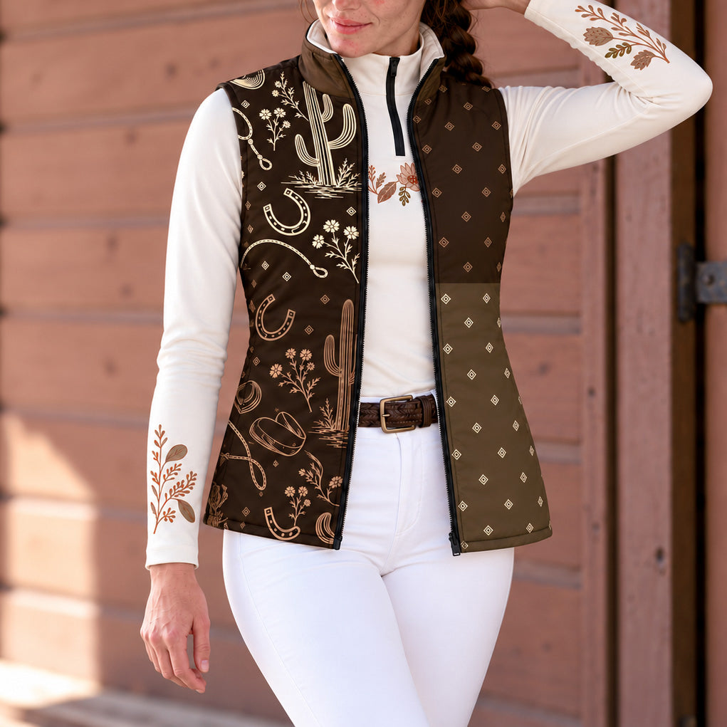 Woman wearing a brown reversible quilted vest with western artwork over a white quarter zip top and white riding pants in front of brown wooden siding