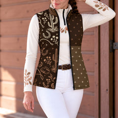 Woman wearing a brown reversible quilted vest with western artwork over a white quarter zip top and white riding pants in front of brown wooden siding