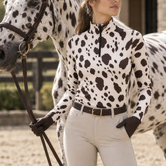 Woman wearing the spotted long sleeve quarter zip riding top with breeches, leather belt and riding gloves beside a spotted Appaloosa horse