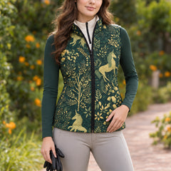 Model wearing the reversible quilted equestrian vest with the dark green unicorn and foliage print side facing forward over a dark green and white quarter zip top, with light gray riding breeches and black riding gloves near wooden fencing and orange flowering plants