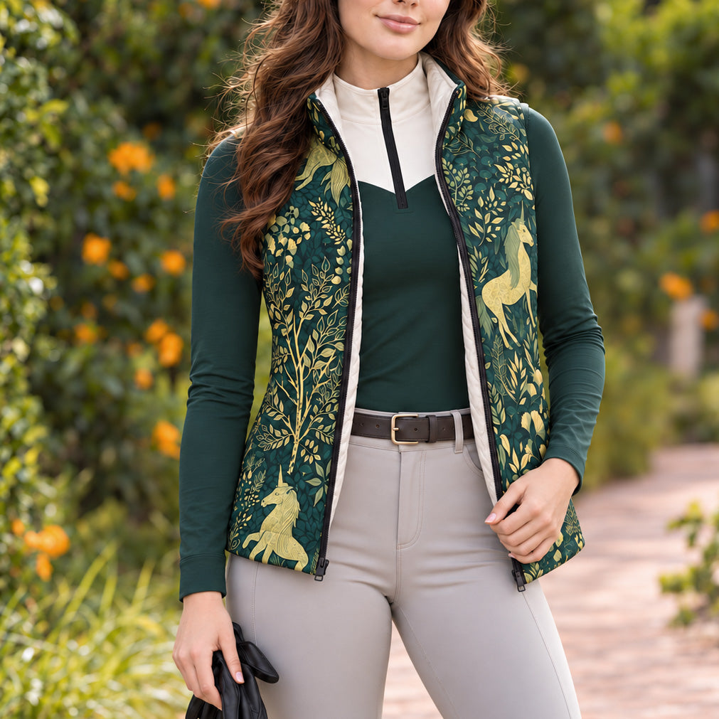 Model wearing the reversible quilted equestrian vest with the dark green unicorn and foliage print side facing forward over a dark green and white quarter zip top, with light gray riding breeches and black riding gloves near wooden fencing