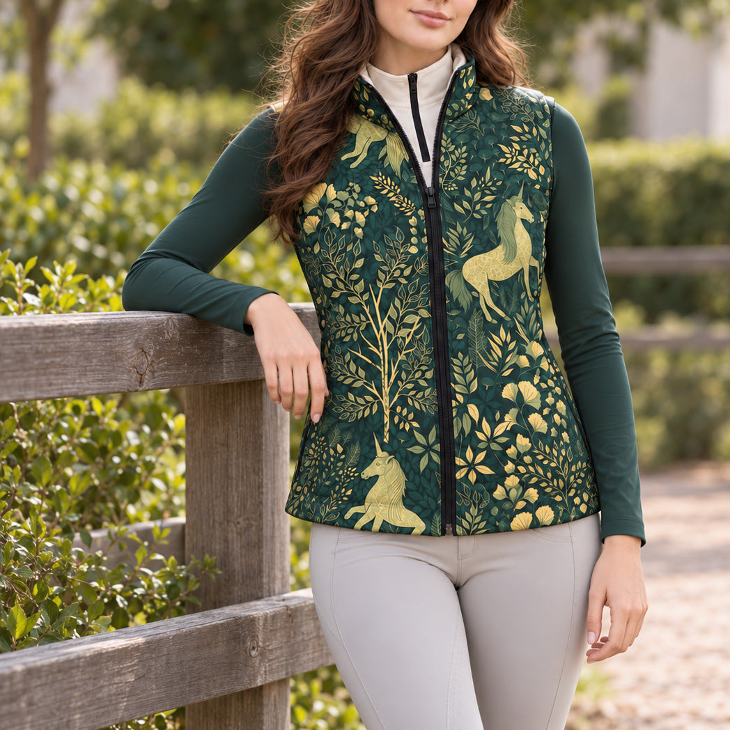 Model wearing the reversible quilted equestrian vest with the dark green unicorn and foliage print side facing forward over a dark green and white quarter zip top, with light gray riding breeches and black riding gloves near wooden fencing and green foliage