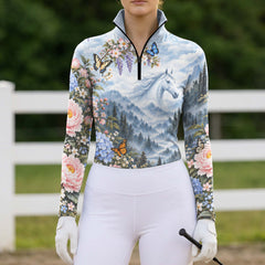 Model in the printed long sleeve quarter zip riding top with white riding breeches, holding a black equestrian helmet and black riding crop while wearing white riding gloves in an arena setting